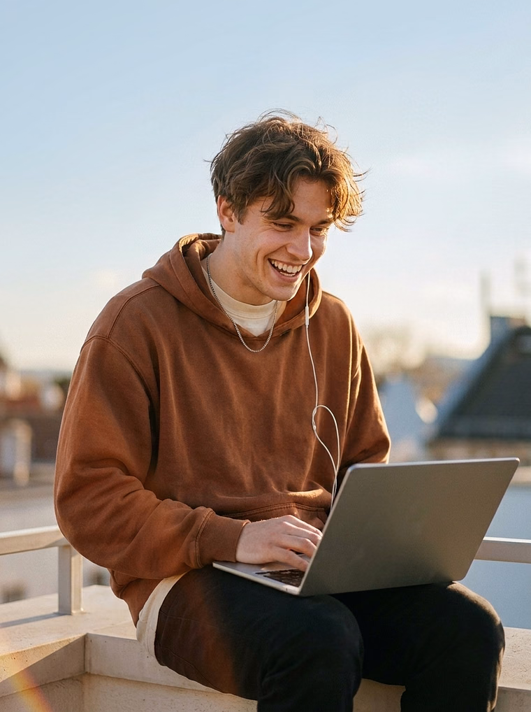 Young developer grinning at his laptop on a rooftop in warm afternoon light, chatting with his AI assistant