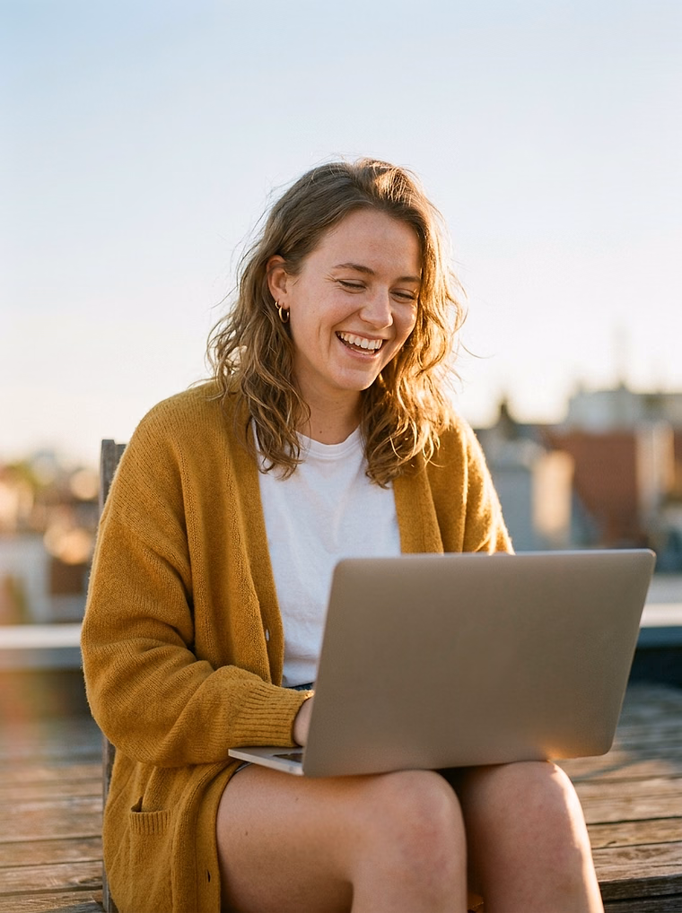 Young woman with wavy hair in a mustard cardigan laughing at her laptop on a rooftop at golden hour