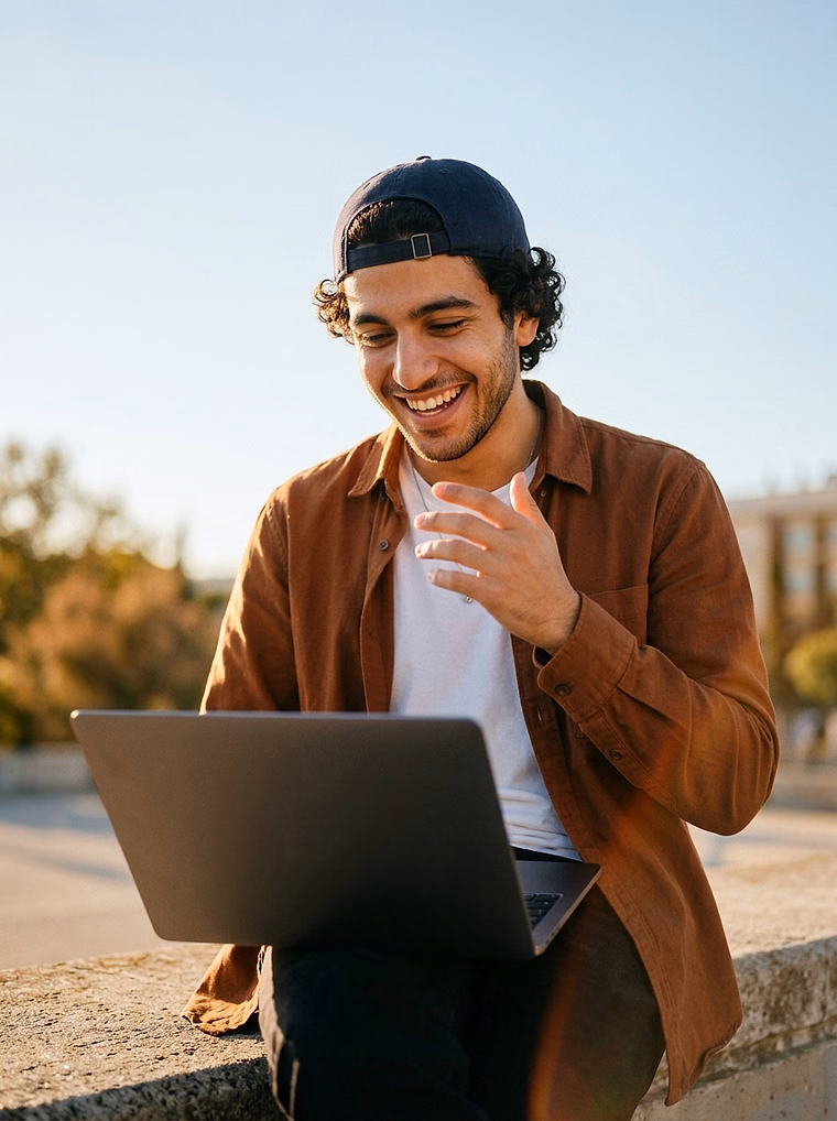 Young man in a backwards cap grinning at his laptop on a low wall in golden light