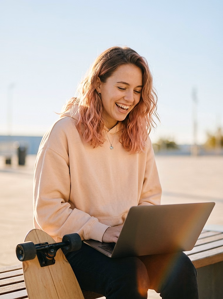 Young woman with pink-tinted hair laughing at her laptop beside a skateboard